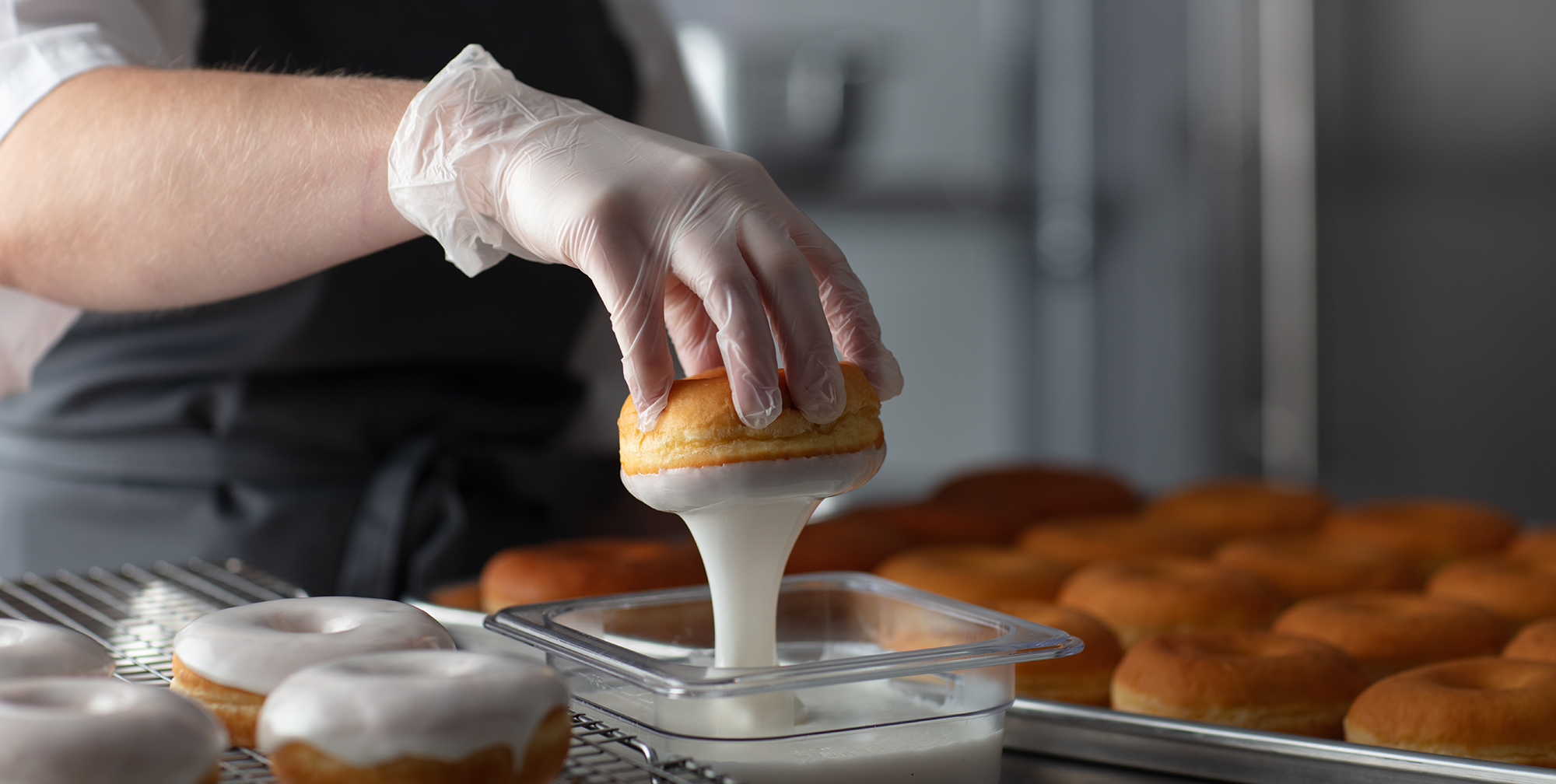 Baker dipping donut in icing with a sheet of donuts in the background