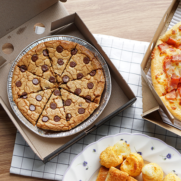 Chocolate chip cookie cake in a box next to pizza