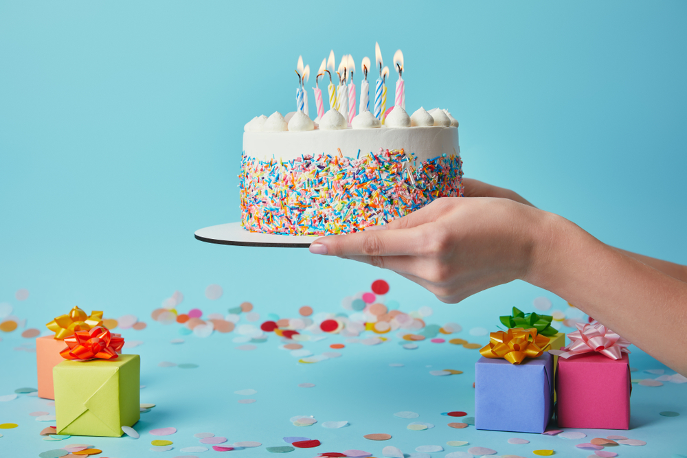 Hands holding a finished decorated birthday cake on a cake pad with small presents and confetti in the background