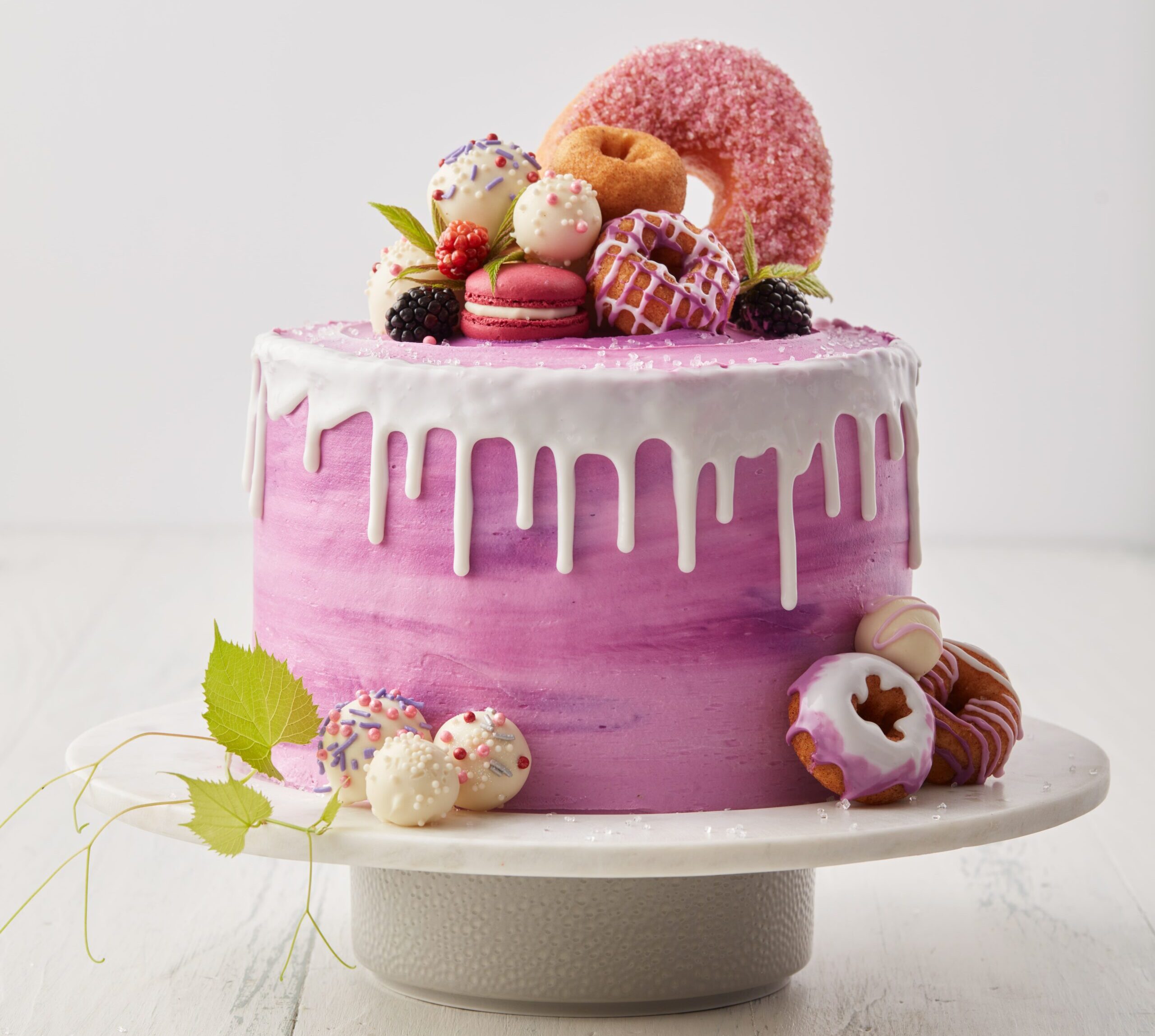 purple frosted cake decorated with donuts, macaroons and white dripping frosting on a cake stand